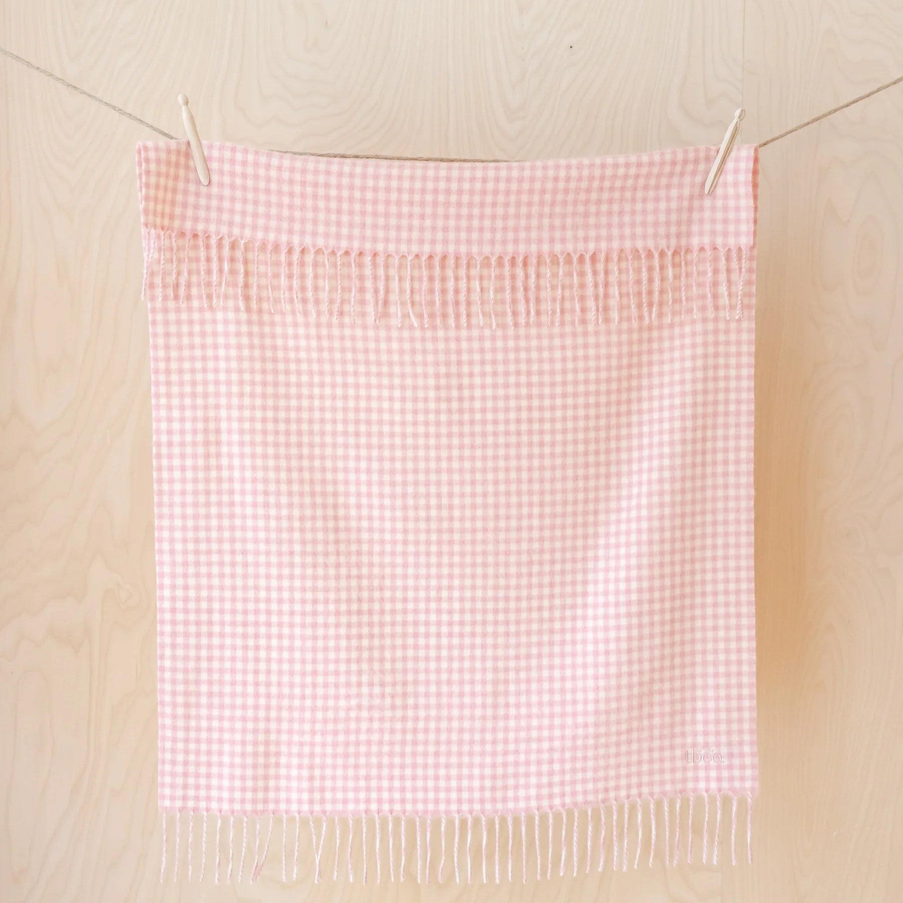 A light pink and white gingham checkered towel with fringed edges hanging from clips on a string against a light wood-paneled wall.